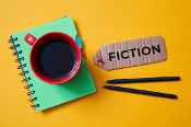Overhead view of a notepad, a mug of coffee, two bens, and a cardboard tag that says "fiction"
