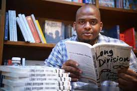 Author Kiese Laymon holding his novel, Long Division, and starting at the camera